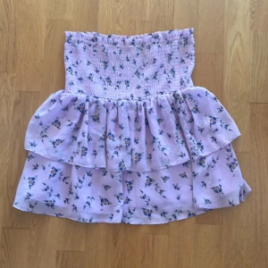 Ljuslila blommig volangkjol - Supersöt ljuslila kjol med blommigt mönster och volanger. Kjolen har smock i midjan för en bekväm passform och är perfekt för varma dagar. Den är kort och har en luftig känsla.