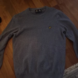 Grå stickad tröja från Lyle & Scott - Säljer en klassisk grå stickad tröja från Lyle & Scott med rund halsringning och den ikoniska gula loggan broderad på bröstet. Perfekt för lager-på-lager och passar till det mesta. Snygg och enkel design!