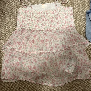 Blommig sommarklänning med volang - Söt blommig klänning med smock upptill och tunna axelband. Klänningen har dubbla volanger och ett luftigt, lätt tyg i vitt med rosa och gröna blommor. Perfekt för varma dagar och en romantisk look. skriv privat för fler frågor eller bilder, kan även gå ner i pris💞