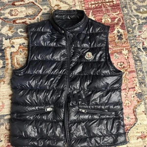Svart dunväst från Moncler - Äkta svart dunväst från Moncler med hög krage och dragkedja framtill. Västen har två fickor med dragkedja och Moncler-logga på bröstet. Perfekt för lager-på-lager och stilren look. 