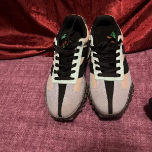 New Balance sneakers i flera färger - Säljer ett par New Balance sneakers med svart, ljusblå, beige och orange detaljer. Skorna har svart snörning, rund tå och platt sula. Perfekta för dig som gillar en sportig och trendig stil.