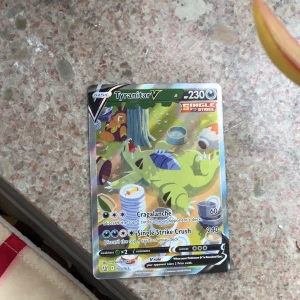 Pokémonkort Tyranitar V - Säljer ett Pokémonkort av Tyranitar V från Single Strike-serien. Kortet har färgstark illustration där Tyranitar ligger bland matrester. HP 230, attacker Cragalanche och Single Strike Crush. Perfekt för samlare eller Pokémon-fans!