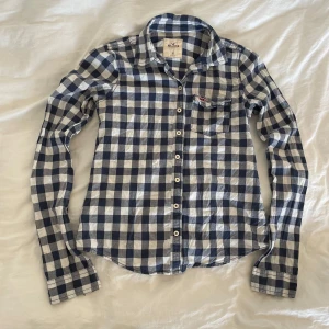 Hollister Blårutig skjorta - Säljer en klassisk blårutig skjorta från Hollister med vit och marinblå färg. Skjortan har lång ärm, knappar framtill och en bröstficka med Hollisters logga broderad - Jag har 20% bundle rabatt om du köper 2 plagg 🦋