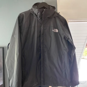 Svart vindjacka från The North Face - Säljer en svart vindjacka från The North Face med huva och dragkedja. Jackan har logga på bröstet och ryggen, samt justerbar huva. Passar även M