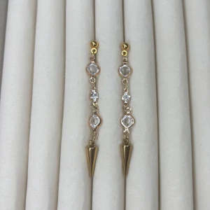 Långa guldörhängen med kristaller och spetsig detalj - Säljer ett par eleganta, långa örhängen i guld med tre klara kristaller på rad och en spetsig, konformad detalj längst ner. Perfekta för att ge en extra touch till din outfit.