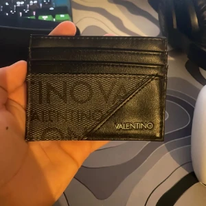 Svart korthållare från Valentino - Snygg svart korthållare från Valentino med stilren design. Framsidan har både slät och mönstrad yta samt guldfärgad logotyp. Perfekt för kort och enkel att bära med sig.