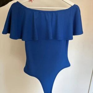 Blå offshoulder body med volang - Säljer en stilren blå body med bred volang upptill som ger en trendig off-shoulder-look. Bodyn har en enkel och elegant design utan synliga mönster eller detaljer. Perfekt för en utekväll eller annan tillställning. 