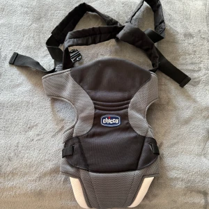 Svart och grå bärsele från Chicco - Säljer en svart och grå bärsele från Chicco med justerbara remmar och vadderad insida för extra komfort. Passar barn från 3,5 kg till 9 kg. Perfekt för att bära barnet nära kroppen och ha händerna fria.