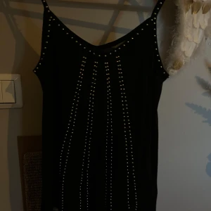 Svart linne med nitar från Lindex - Säljer ett svart linne  från Lindex med smala axelband och dekorativa silverfärgade pärlor i mönster framtill. Linnet har en enkel, rak passform och är ärmlös. Perfekt för dig som gillar en stilren look med lite edge.