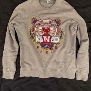 Grå sweatshirt med tigerbroderi från Kenzo - Säljer en grå sweatshirt från Kenzo med ikoniskt tigerbroderi och färgglada detaljer framtill. Storlek xs men lite liten i storleken. Ny pris på denna är 2500:- i och med att den är lite sliten så säljer jag den för 500!