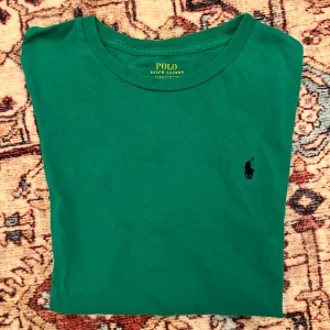 Grön t-shirt från Polo Ralph Lauren - Snygg grön t-shirt från Polo Ralph Lauren med klassisk rund hals och broderad marinblå logga på bröstet. Perfekt för en stilren och avslappnad look. Tillverkad i mjuk bomull för extra komfort.