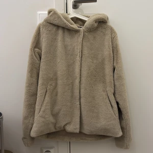 Beige teddy/päls med huva - Mysig beige teddy/pälsjacka från Kappahl med huva och dragkedja framtill. Jackan har två fickor på sidorna och är perfekt för kyligare dagar. Den fluffiga ytan ger en trendig och avslappnad look.