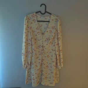 Blommig gul klänning från Nelly x Glamorous - Söt, gul och en kortare klänning med blommigt mönster i orange och lila från Nelly x Glamorous. Klänningen har v-ringning, knappar framtill och långa ärmar. Perfekt inför festliga tillfällen i sommar😍
