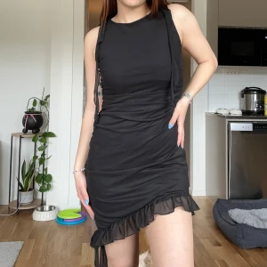 Svart kort klänning med volang från NA-KD - Säljer en svart kort klänning från NA-KD med volangkant nertill och knytband på axlarna. Klänningen har en tight passform och är ärmlös, perfekt för dig som gillar en stilren look med lite extra detalj. Aldrig använd 