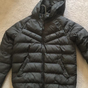 Svart dunjacka från Nike med huva - Säljer en svart dunjacka från Nike i storlek XL. Jackan har huva, dragkedja framtill och två fickor med knappar. Klassisk pufferstil med diskret Nike-logga på bröstet. Perfekt för kyliga dagar.