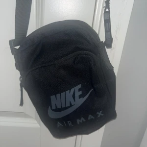 Svart axelväska från Nike Air Max - Säljer en svart axelväska från Nike Air Max med justerbar rem och dragkedja. Väskan har ett stort huvudfack och en mindre ficka framtill, perfekt för att bära med sig det viktigaste. Stor Nike-logga på framsidan.