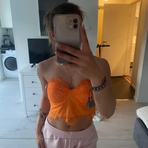 Orange spetsig croppad bustier - Säljer en orange croppad bustier med spetsdetaljer och bygel. Toppen är ärmlös och har dragkedja i ryggen. Perfekt för att ge en färgklick till outfiten.