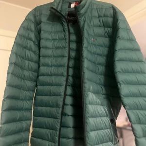 Grön pufferjacka från Tommy Hilfiger - Snygg grön pufferjacka från Tommy Hilfiger med svart dragkedja och diskret logga på bröstet. Jackan har quiltad design och klassisk passform. Enkel att matcha med olika outfits. Har aldrig använts  ORD PRIS: 2299