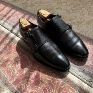 Svarta dubbla munkstrap-skor i skinn - Säljer ett par svarta dressade munkstrap-skor från Excalibur. Skorna har dubbla spännen i silver, rund tå och är tillverkade i slätt och präglat skinn. Perfekta för dig som vill ha en stilren och klassisk look.