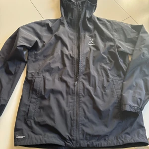 Svart vindjacka från Haglöfs - Säljer en svart vindjacka från Haglöfs med dragkedja framtill och hög krage. Jackan har två sidofickor och är tillverkad i ett lätt syntetmaterial, perfekt för blåsiga dagar. Klassisk och stilren design med diskret logga på bröstet. Stl L dam men passar även M herr.