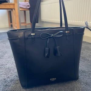 Svart handväska från Guess  - Snygg svart handväska från Guess med dekorativ rosett framtill och guldfärgade detaljer. Väskan har dragkedja upptill, innerfack och ett ytterfack med dragkedja. Insidan är ljus med Guess-logga. Perfekt storlek för vardagens alla prylar. Inga defekter. Använt fåtal gånger. 