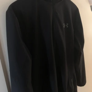 Svart Windbreaker från Under Armour - Säljer en svart Windbreaker från Under Armour med dragkedja och huva. Storlek L. I JD ny pris 1200 säljer för 750kr