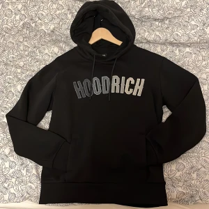 Svart hoodie från Hoodrich - Säljer en svart hoodie från Hoodrich i storlek M. Tröjan har huva med dragsko och en stor logga i vitt och grått över bröstet. Perfekt för en avslappnad och cool stil.