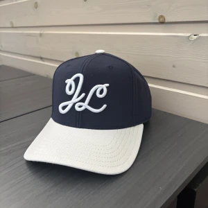 Marinblå och vit keps från J.Lindeberg - Snygg marinblå keps från J.Lindeberg med vit böjd skärm och broderad JL-logga framtill. Justerbar snapback baktill för perfekt passform. Klassisk och stilren design med grönt under skärmen. Aldrig använd 