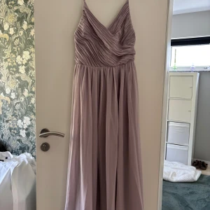 Ljuslila långklänning med plisserad överdel - Säljer en elegant ljuslila långklänning för 500kr med smala axelband och v-ringad, plisserad överdel. Klänningen har ett flytande fall och markerad midja, perfekt för bal eller fest. Materialet känns mjukt och ger en lyxig känsla. Klänningen är endast använd en kväll under studentbalen och är precis som ny.