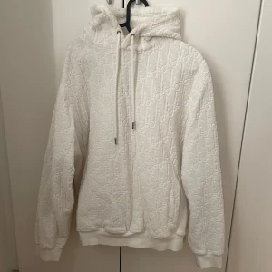 Vit hoodie med broderat mönster - Säljer en vit hoodie med broderat mönster och huva. Tröjan har dragsko i halsen och ribbade muddar vid ärmslut och nederkant. Perfekt för en stilren och bekväm look.
