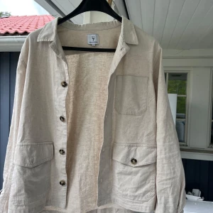 Beige linneskjorta jacka  från Urban Pioneers - Säljer en stilren beige skjorta från Urban Pioneers i linne. Skjortan har tre fickor framtill, knappar och klassisk krage. Perfekt för dig som gillar en avslappnad men snygg look. Passar till många olika tillfällen!