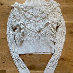 Vit stickad tröja med volang från Zara - Supersöt vit stickad tröja från Zara med höga volangdetaljer och strukturstickat mönster. Tröjan har lång ärm och hög hals, perfekt för att lyfta din outfit med en romantisk touch.