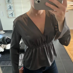 Mörkgrå omlottblus med v-ringning - Snygg mörkgrå omlottblus med v-ringning och långa ärmar. Blusen har resår i midjan som ger en fin form och är tillverkad i ett glansigt material. Perfekt att styla med jeans eller kjol.