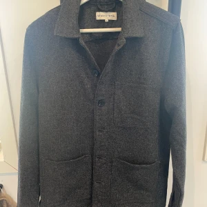 Grå overshirt från Studio Total - Säljer en stilren grå overshirt från Studio Total. Jackan har klassisk krage, knäppning framtill med knappar och tre praktiska fickor. Perfekt att slänga över en t-shirt eller hoodie för en clean look.