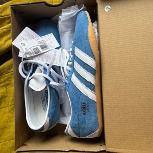 Adidas Tokyo W blå sneakers - Säljer ett par Adidas Tokyo W sneakers i blå mocka med vita ränder och vit snörning. Skorna har en klassisk retrostil med gummisula och rund tå. Perfekta för dig som gillar sportig och stilren look.