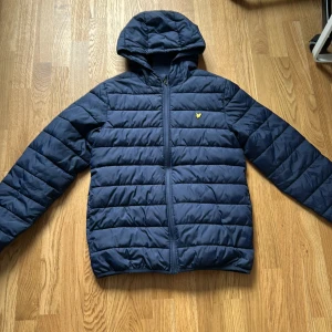 Blå pufferjacka från Lyle & Scott Junior - Säljer en blå pufferjacka från Lyle & Scott Junior med huva och dragkedja framtill. Jackan har ett quiltat mönster och den klassiska gula loggan på bröstet. Perfekt för kyligare dagar och riktigt skön att ha på sig.