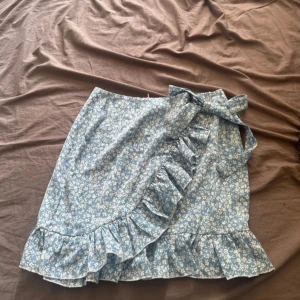 Blommig omlottkjol med volang och rosett - Supersöt ljusblå omlottkjol med vitt blommönster. Kjolen har volang längs kanten och en stor rosett i midjan. Perfekt för dig som gillar romantisk och feminin stil.