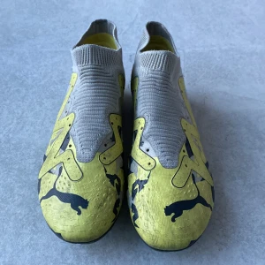 Puma fotbollsskor i grått och gult - Säljer ett par grå och gula Puma fotbollsskor med svart logga och mönster. Skorna har en strumpliknande ovandel utan snörning och platt sula med dobbar för bra grepp på planen.