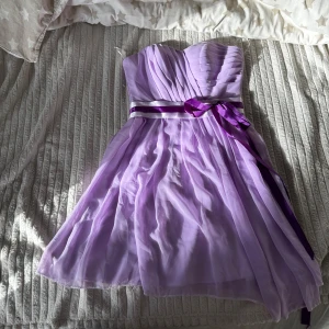 Ljuslila axelbandslös klänning med rosett - Söt ljuslila axelbandslös klänning med draperad överdel och markerad midja med lila och vit satinband samt en dekorativ rosett. Kjolen är lätt utställd och har flera lager för extra volym. Perfekt för bal eller fest.