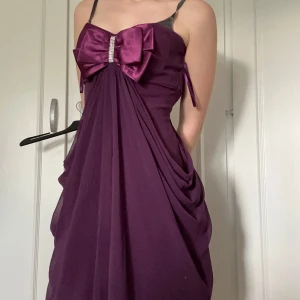 Lila kortklänning med rosett - Supersöt lila kortklänning med tunna axelband, stor satinrosett framtill och strassdetalj i mitten. Klänningen har ett luftigt fall och är perfekt för speciella tillfällen. Materialet känns lätt och glansigt.