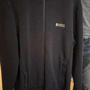 Svart zipjacka från BOSS, storlek L - Snygg svart zipjacka från BOSS med stilren design och logga på bröstet. Jackan har hög krage, hel dragkedja och två fickor framtill. Perfekt för en clean och sportig look. Materialet är mjukt och skönt, passar till många outfits.endast använd 2 gånger så gott som ny!