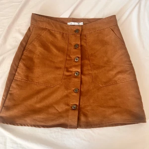 Brun mockakjol med knappar - Snygg brun kortkjol i mocka-look med knappar framtill och två stora fickor. Kjolen har en A-linjeformad passform och passar perfekt till en cool vardagsstil. Enkel att matcha med stickade tröjor eller t-shirts.