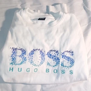 Vit sweatshirt från Hugo Boss - Vit sweatshirt från Hugo Boss med stort BOSS-tryck framtill i blå och turkosa toner. Tröjan har rund hals, långa ärmar och ribbade muddar. Snygg och clean design med digitalt inspirerat mönster på loggan. Perfekt för dig som gillar stilrena märkesplagg.