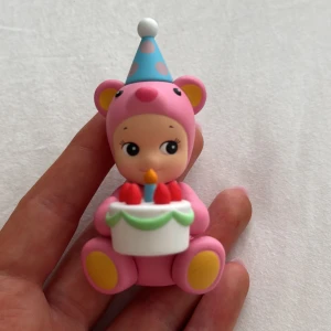 Sonny Angel - Bilderna visar en söt figur i rosa björndräkt med födelsedagshatt och tårta, men ingen bok syns. Ingen information om titel, författare eller bokinnehåll kan utläsas.