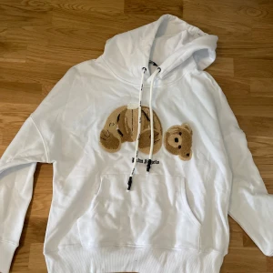 Vit Palm Angels hoodie med nallebrodyr - Vit hoodie från Palm Angels med stor broderad nalle på bröstet. Tröjan har huva med snörning där Palm Angels-loggan är tryckt på snörena och en klassisk känguruficka framtill. Oversized passform och mjukt bomullsmaterial gör den både snygg och bekväm. Den är i storlek M och helt oanvänd.