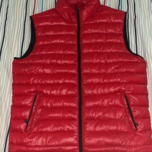 Röd dunväst från Raw Blue 2XL - Röd dunväst från Raw Blue i storlek 2XL. Västen har quiltad design, hög krage och svarta dragkedjor både framtill och vid fickorna. Perfekt för lager-på-lager och ger en sportig vibe. Materialet är glansigt och lätt, vilket gör den smidig att bära.