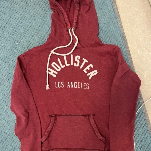 Vinröd hoodie från Hollister - Säljer en vinröd hoodie från Hollister med vit text 'Hollister Los Angeles' på bröstet. Tröjan har huva med snörning, magficka och långa ärmar med ribbade muddar och tumhål.