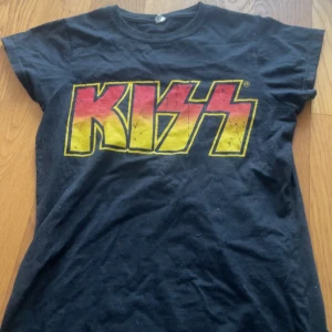 Svart KISS t-shirt med tryck - Cool svart t-shirt med stort KISS-tryck i gult och rött på bröstet. Klassisk rund hals och korta ärmar. Perfekt för dig som gillar rock och vill sticka ut med en ikonisk bandtröja. Materialet känns mjukt och skönt mot huden.