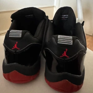 Nike Air Jordan 11 Low Bred svart/röd - Säljer ett par Nike Air Jordan 11 Low Bred med svart ovandel i mesh och lackdetaljer, samt röd sula. Klassisk Jumpman-logga i rött på hälen. Skorna har snörning och en låg siluett som ger en sportig vibe.