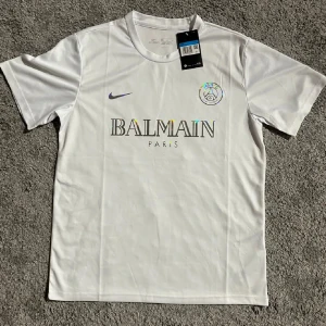 Vit PSG x Balmain fotbollströja Nike - Vit fotbollströja från Nike med Paris Saint-Germain och Balmain Paris tryck på bröstet. Klassisk rund hals, korta ärmar och lätt, andningsbart material. Snyggt holografiskt tryck på loggan och texten. Perfekt för dig som gillar exklusiva samarbeten och stilren design.